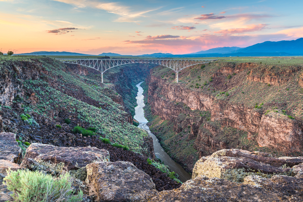 a very western landscape. a rocky gorge with a light spray of green plants on top. a metal bridge spans the gorge and we can see the river flowing into the distance