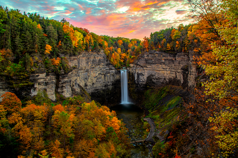 stunning waterfall coming down a rocky gorge. atop the gorge are many trees, most of which bear autumn foliage. a colorful sunset in the top