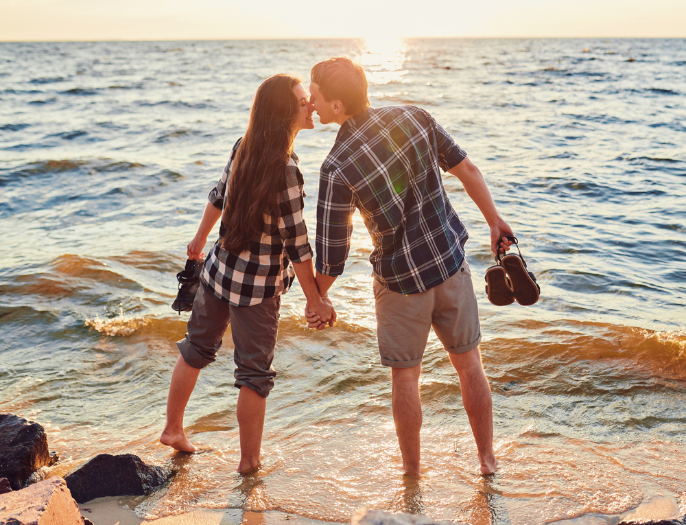 couple holding hands and standing in the waves, kissing in front of sunset on a large lake | romantic getaways in michigan