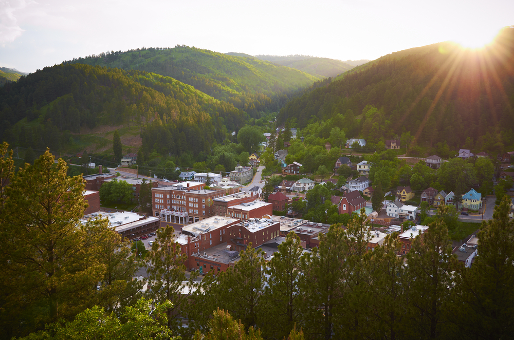 an overhead view of deadwood south dakota. red bricked buildings in a valley surrounded by lush green hills. the sun is shining from the right onto the town
