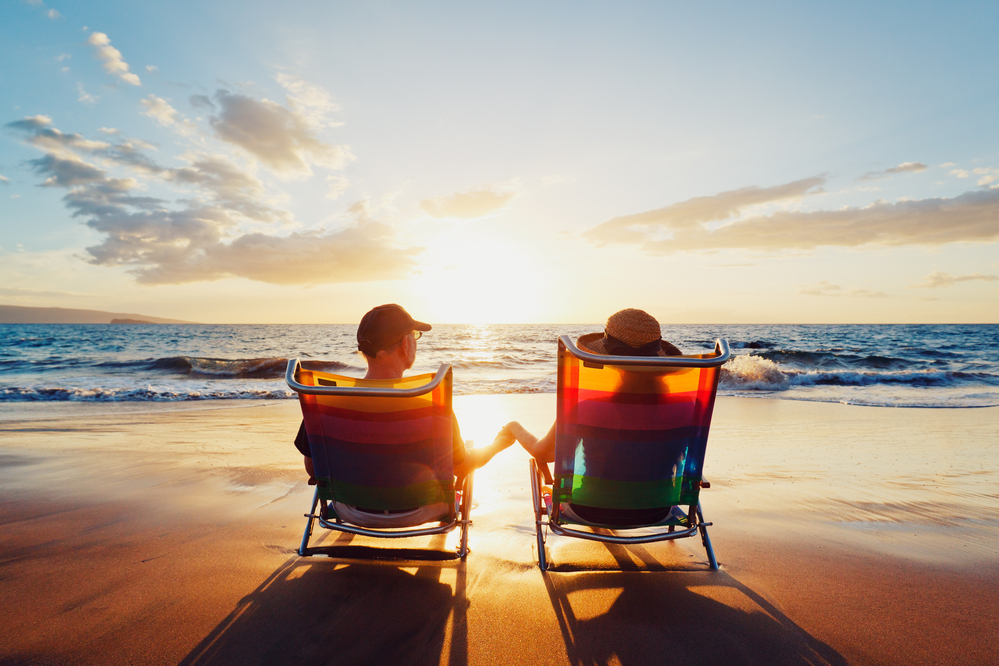 a couple sits in chairs on the beach. they are facing the ocean and holding hands. the sun is almost setting over the water.